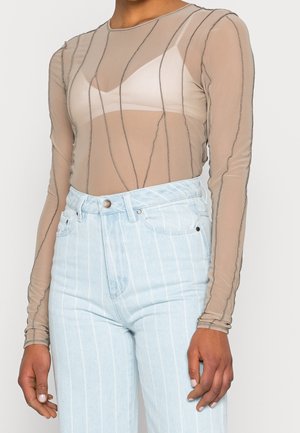 Personne portant un haut beige transparent à manches longues avec des lignes noires ondulées et un soutien-gorge blanc en dessous, associé à un jean taille haute bleu clair à rayures fines.