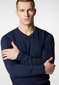 Granatowy sweter w serek o prążkowanej teksturze. Długie rękawy z dopasowanymi mankietami, noszony przez modela z założonymi rękami. Neutralne tło.