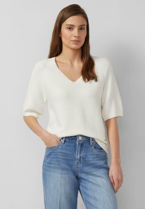 Donna con capelli castani lunghi che indossa un maglione bianco a scollo a V con maniche corte e jeans blu a vita alta, con una mano in tasca.