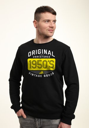 Ung mand med kort hår iført sort sweatshirt med teksten "Original and Unrestored 1950's Vintage Goods" og gul grafik, der kigger sidelæns.