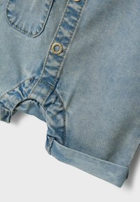 Romper de denim azul claro con botones en la parte frontal, aberturas de pierna con volantes y dobladillos enrollados. Presenta costuras doradas y un bolsillo delantero.