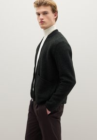 Cardigan verde scuro con cerniera e finitura texture, polsini a costine e due tasche anteriori, abbinato a pantaloni marroni e dolcevita bianca.