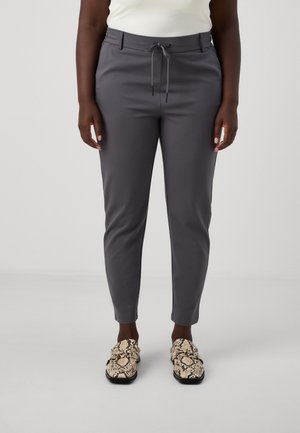 Person wearing fitted dark gray pants with a front drawstring and beige snake-print slip-on shoes, standing against a plain light background.