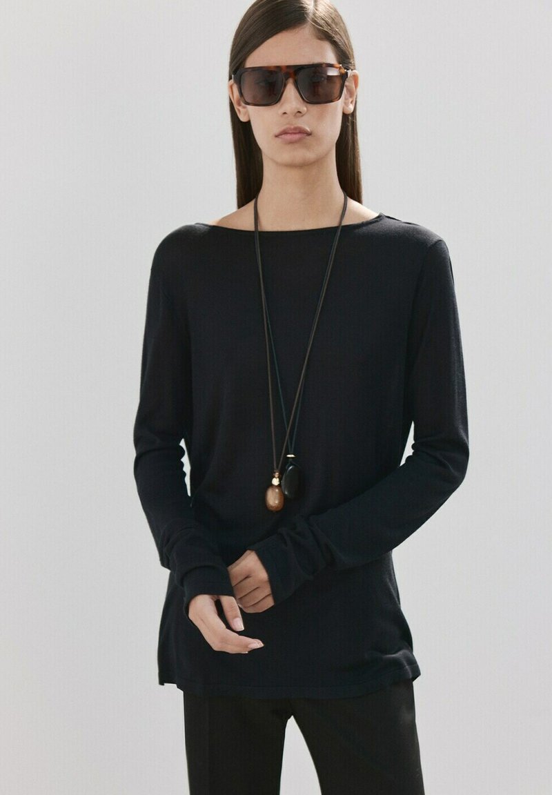 Frau mit übergroßen dunklen Sonnenbrillen, einem schwarzen Langarmshirt und zwei Anhänger-Halsketten vor einem schlichten hellen Hintergrund.