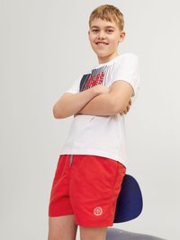 T-shirt grafica bianca con motivi blu e rossi, abbinata a pantaloni corti rossi con logo di basket. Tessuto morbido, vestibilità casual.