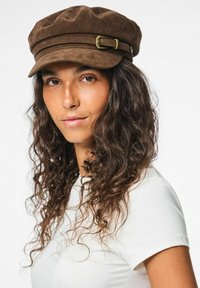 Gorra de ante marrón con una parte superior redondeada, dos bandas horizontales y un detalle decorativo de hebilla dorada en el lateral. Se lleva con una camisa blanca.