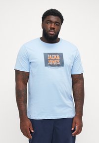 Ανοιχτό μπλε βαμβακερό T-shirt με κοντά μανίκια, που διαθέτει τετράγωνο γραφικό που γράφει "JACK & JONES" σε πορτοκαλί και μαύρο, με λεία υφή.