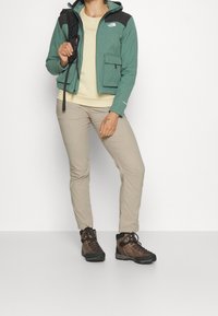 Grön fleecetröja med svarta detaljer, beige skjorta, tanbyxor och bruna vandringskängor. Inkluderar dragkedjefickor och huva.