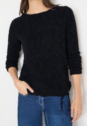 Femme portant un pull noir duveteux à manches longues avec un cordon latéral et un jean bleu, main dans la poche, debout devant un fond blanc.