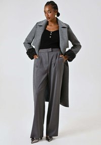 Cappotto di lana grigio con dettagli in pelliccia ai polsini, abbinato a pantaloni grigi a vita alta. La maglia nera a costine presenta dettagli con bottoni.