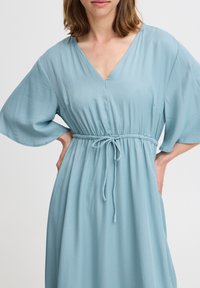Helles blaues Kleid mit V-Ausschnitt, kurzen weiten Ärmeln und elastischem Taillenbund. Es verfügt über einen Kordelzug in der Taille und einen weichen, glatten Stoff.