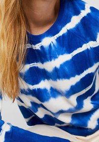 Sinivalkoinen pilkullinen collegepaita ribbineuloksella, jossa on pyöreä kaulus ja pitkät hihat, ja aaltoileva kuvio.