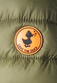 Olivgrüner Pufferjackenstoff mit einem runden orangefarbenen Gummilogo, das einen Enten und den Schriftzug "SAVE THE DUCK" zeigt. Texturierte und lebendige Details.