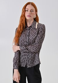 Donna dai capelli rossi che indossa una camicia nera a bottoni con fantasia e pantaloni neri, in piedi con le braccia incrociate su uno sfondo chiaro.