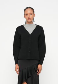 Černý cardigan s V-výstřihem, zapínáním na knoflíky a dlouhými rukávy, zkombinovaný se šedým topem s kulatým výstřihem a černou sukní s volány.