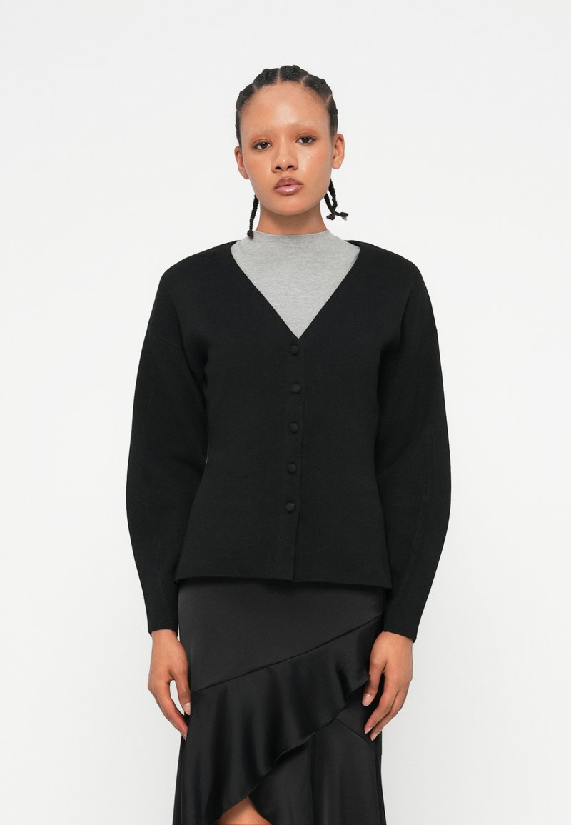 Černý cardigan s V-výstřihem, zapínáním na knoflíky a dlouhými rukávy, zkombinovaný se šedým topem s kulatým výstřihem a černou sukní s volány.