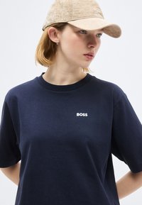 Νεαρή γυναίκα που φοράει μπεζ καπέλο και μπλε ναυτικό T-shirt με το λογότυπο "BOSS", κοιτάζοντας πλάγια σε λευκό, απλό φόντο.