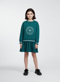 Teal sweatshirtklänning med långa ärmar, med en cirkulär grafik på bröstet och en insydd kjol. Till denna är den matchad med svarta skor.