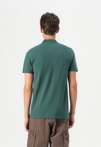 Zelené bavlněné polo tričko s volným střihem, krátkými rukávy a límečkem, zkombinované s hnědými cargo šortkami. Hladká textura a minimální detaily.