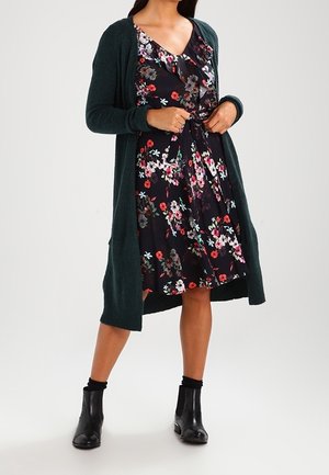Femme ajustant la ceinture taille sur une robe noire à motifs floraux mi-genoux, portant un long cardigan vert foncé et des bottines noires.