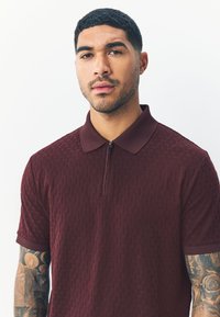 Polo color bordeaux con motivo strutturato, maniche corte e chiusura con zip. Presenta un colletto a tinta unita e dettagli a contrasto.