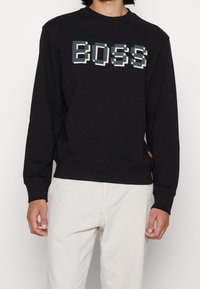 Sudadera negra con letras pixeladas de "BOSS" en azul y blanco. Fabricada con tela suave, con puños y dobladillo acanalados, y pequeña etiqueta de logo.