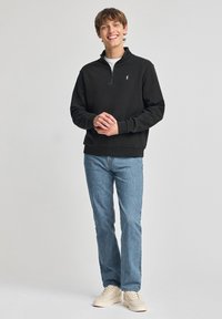 Zwarte zip-up sweatshirt met een staande kraag, gecombineerd met lichtblauwe jeans en beige schoenen. De sweatshirt heeft een klein logo op de linkerborst.