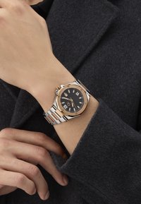 Guld- og sølvtonet ur på håndleddet, har en sort skive med sølvmarker, ottekantet kasse og et segmenteret metalrem.