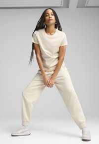 Beige katoenen t-shirt en bijpassende joggers met een subtiel logo. Witte sneakers met beige accenten. Model poseert met gespreide benen.