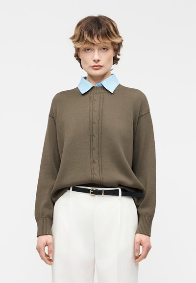 Personne portant un pull marron à torsades sur une chemise bleu clair à col, un pantalon blanc et une ceinture noire, debout devant un fond blanc.