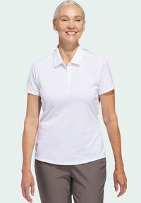 Bílá polo košile s krátkým rukávem, se zapínáním na pět knoflíků. Hladká tkanina a klasický límec, v kombinaci s hnědými kalhotami.
