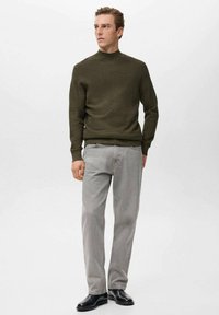 Maglione a maglia verde oliva con colletto alto, abbinato a jeans grigi chiari e scarpe nere. Il modello è in piedi contro uno sfondo bianco semplice.