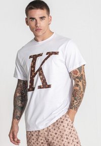 Bílá tričko s velkým hnědým logem "K" s vzorovaným designem. Nosené s vzorovanými béžovými šortkami.