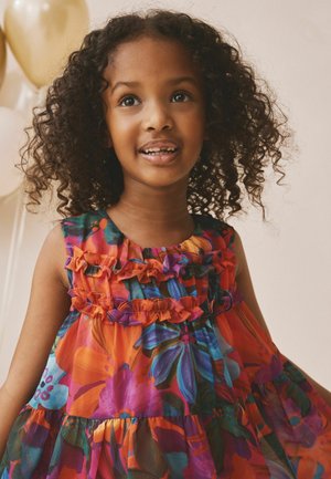 Lächendes junges Mädchen mit lockigem Haar, das ein buntes Blumenkleid mit Rüschen trägt und drinnen steht, im Hintergrund sind Luftballons.