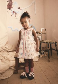 Un vestito senza maniche per bambini presenta un tessuto color crema con stampe di animali colorati, un colletto rotondo e una cerniera frontale rossa. Abbinate calzini rosa e scarpe nere.