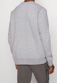 Uomo che indossa un sudario NFL grigio chiaro e pantaloni grigi scuri, visto da dietro su uno sfondo semplice.