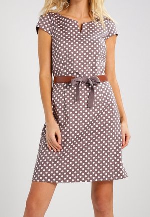 Kobieta ubrana w taupe sukienkę o krótkich rękawach, sięgającą kolan, w białe kropki, z brązowym paskiem związanym w kokardę w talii.
