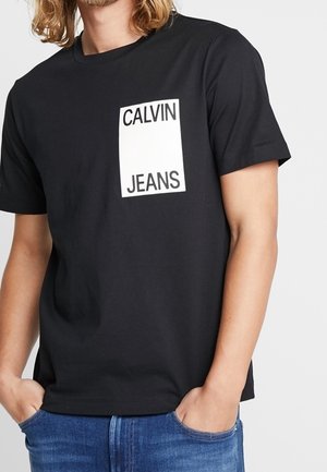 T-shirt en coton noir avec un graphique rectangulaire blanc comportant "CALVIN" au-dessus de "JEANS" sur le côté gauche de la poitrine. Col rond classique.