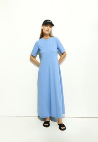 Robe maxi bleu clair à manches courtes, coupe décontractée avec un col rond. Associée à des sandales plates noires et une casquette noire. Tissu lisse.