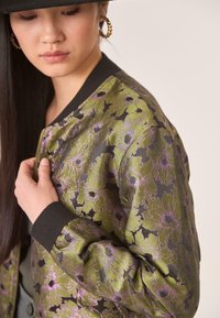 Joven ajustándose una chaqueta bomber con un estampado floral verde y morado, llevando un sombrero negro y pendientes de aro dorados, con un fondo neutro.