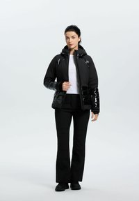 Sort polstret jakke med hætte, med glansfulde detaljer på skuldre og ærmer, parret med sorte bukser og støvler. Glat stofstruktur.