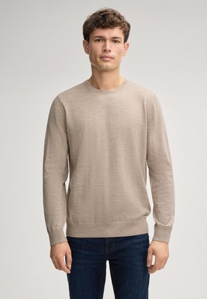 Maglione lungo beige chiaro con maniche lunghe, scollatura tonda e polsini a costine, abbinato a jeans scuri. Texture morbida e design semplice.