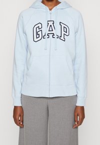 Sudadera con cremallera de color azul claro con bolsillos frontales y el logo "GAP", combinada con pantalones de pierna ancha grises, mostrada desde los hombros hasta las caderas.