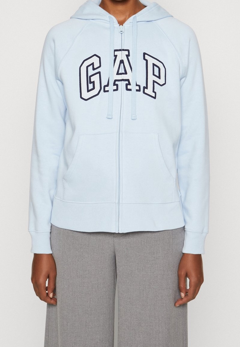 Sudadera con cremallera de color azul claro con bolsillos frontales y el logo "GAP", combinada con pantalones de pierna ancha grises, mostrada desde los hombros hasta las caderas.