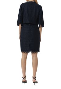 Marineblauwe kanten jurk met een cropped top, voorzien van een hexagonaal patroon en een gegolfde zoom. Enkellaarsjes met enkelbandje maken de outfit compleet.