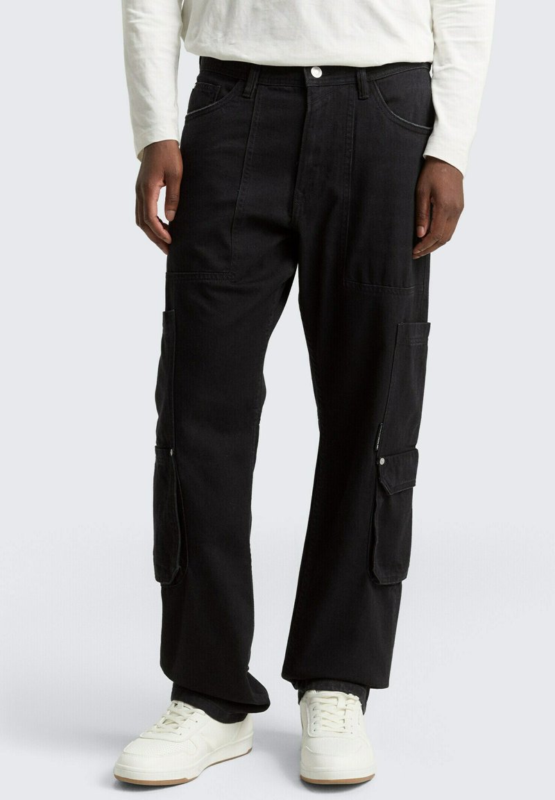Schwarze Cargohosen mit mehreren Taschen, kombiniert mit weißen Sneakern und einem weißen Langarmshirt vor grauem Hintergrund.