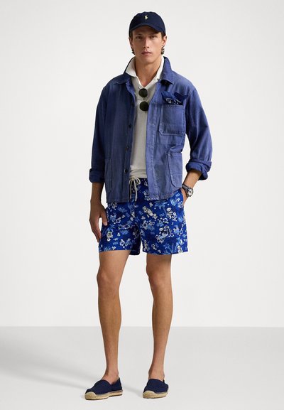 Chaqueta de mezclilla azul, polo blanco, pantalones cortos florales azules y alpargatas marinas. Los accesorios incluyen gafas de sol y una gorra. Look veraniego casual.