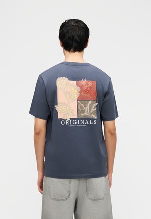 Mężczyzna ubrany w niebieski T-shirt z kwiatowym nadrukiem i tekstem „ORIGINALS designed in Copenhagen” na plecach, w połączeniu z jasnoszarymi szortami.