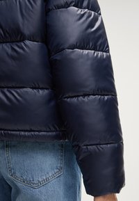 Giacca a vento blu navy con texture liscia e lucida, design trapuntato con ispessimento, e polsini elasticizzati. Indossata con jeans azzurri.
