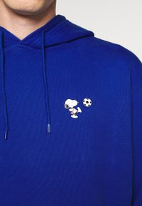 Felpa blu con texture a costine, caratterizzata da un design ricamato di Snoopy che gioca a calcio sulla parte anteriore, con punte del cordino in tono argento.